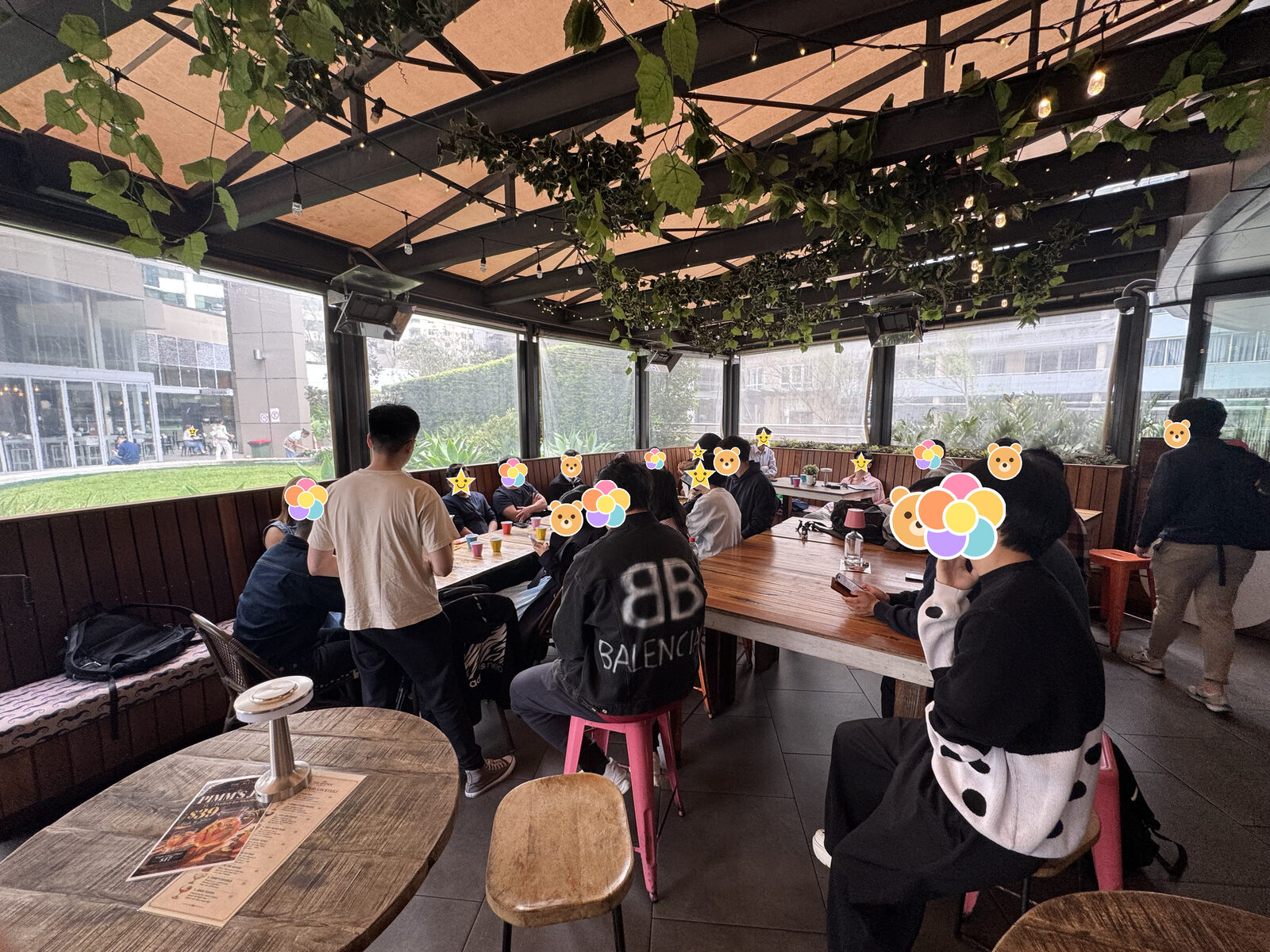 玻璃房的全景,头顶是藤,长桌两边坐满了人,前景是一张圆木桌